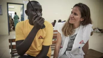 Unsere Mitarbeitenden Noon Makor und Christina Martel arbeiten in dem von uns unterstützen Ameth-Bek-Krankenhauses in Südsudan. Infoabend Mitarbeit: Zwei Mitarbeitende auf Bank, einer mit Radiotelefon