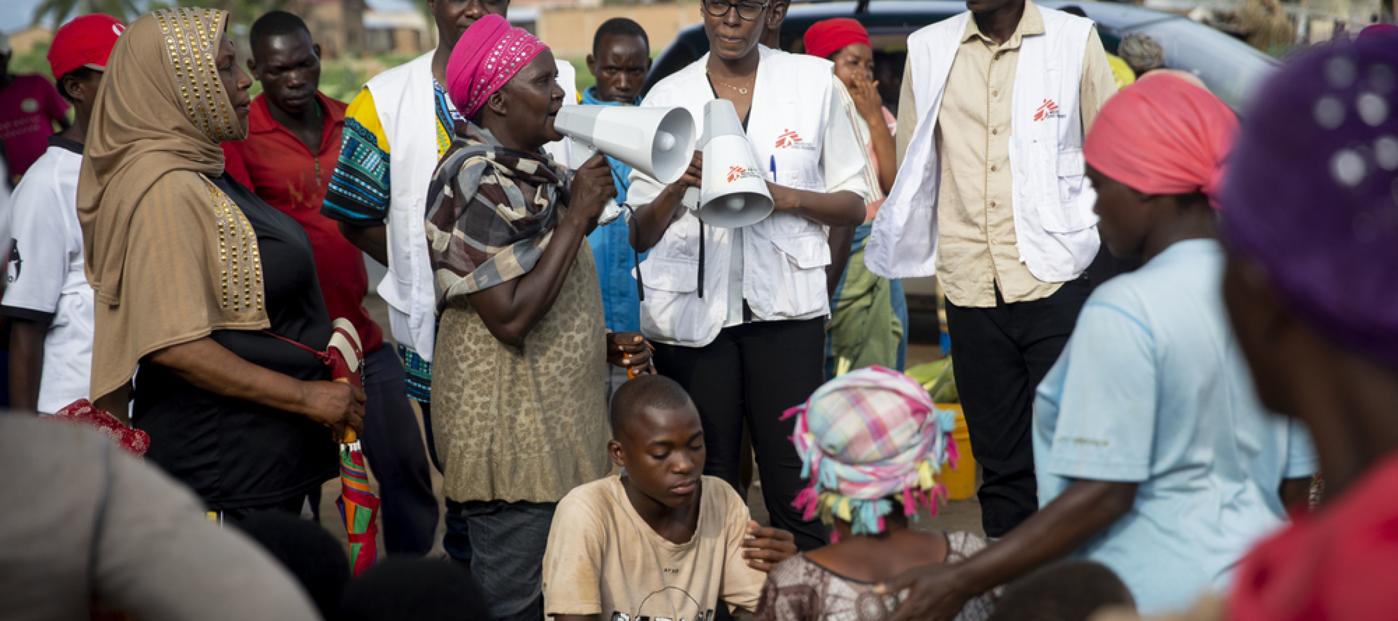 Eine Community Health Mitarbeiterin klärt mit einem Megaphon auf dem Buterere-Markt in Bujumbura über Cholera auf. Um sie herum sind mehrere Mitarbeiter sowie weitere Personen versammelt und hören zu.