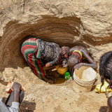 Zwei Frauen stehen in einem Loch im Boden und schöpfen Wasser, andere außenherum sehen zu